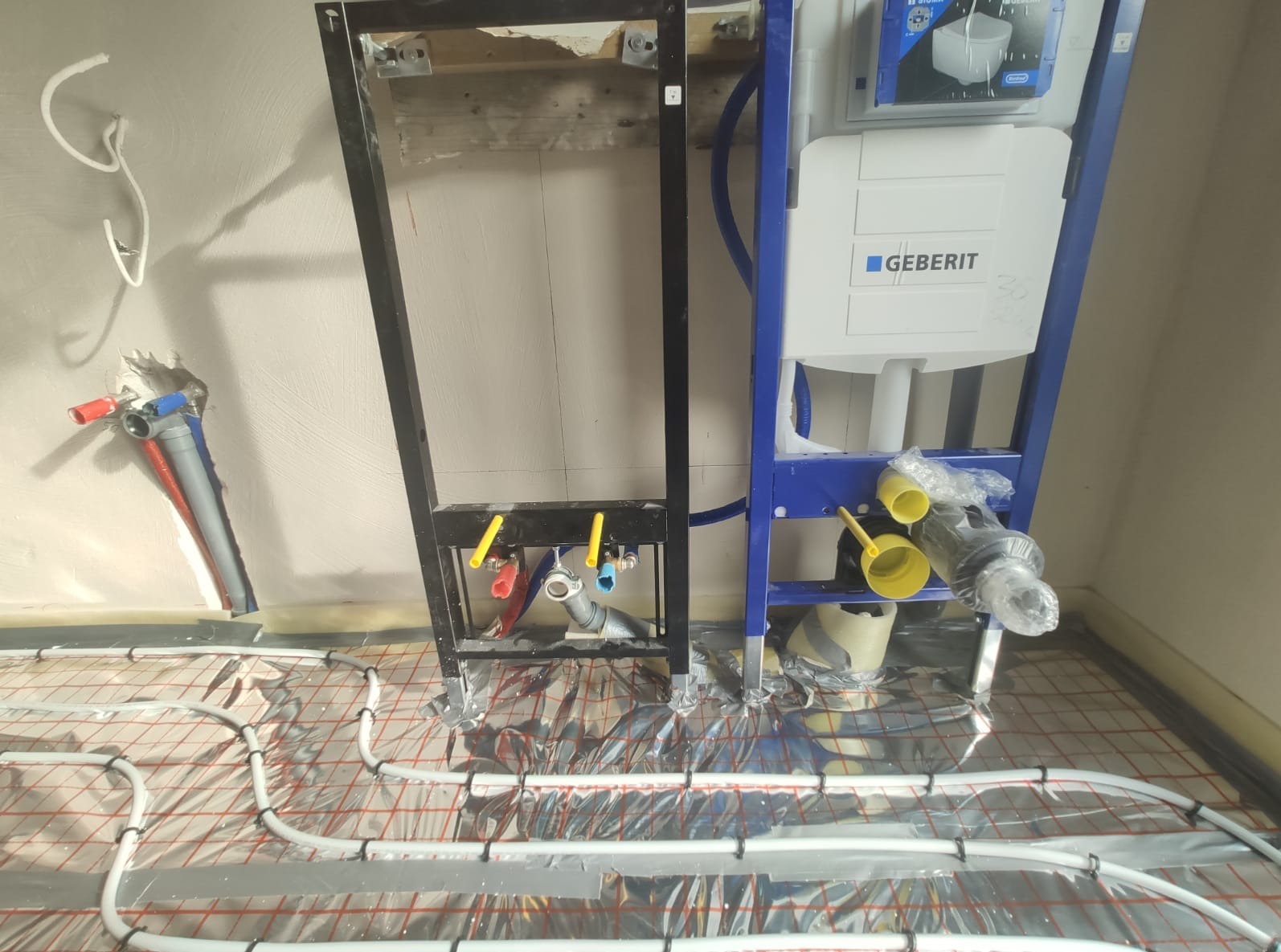 Montaż stelaża podtynkowego Geberit z instalacją ogrzewania podłogowego, widoczne rury doprowadzające wodę, folia aluminiowa pod wężami grzewczymi.