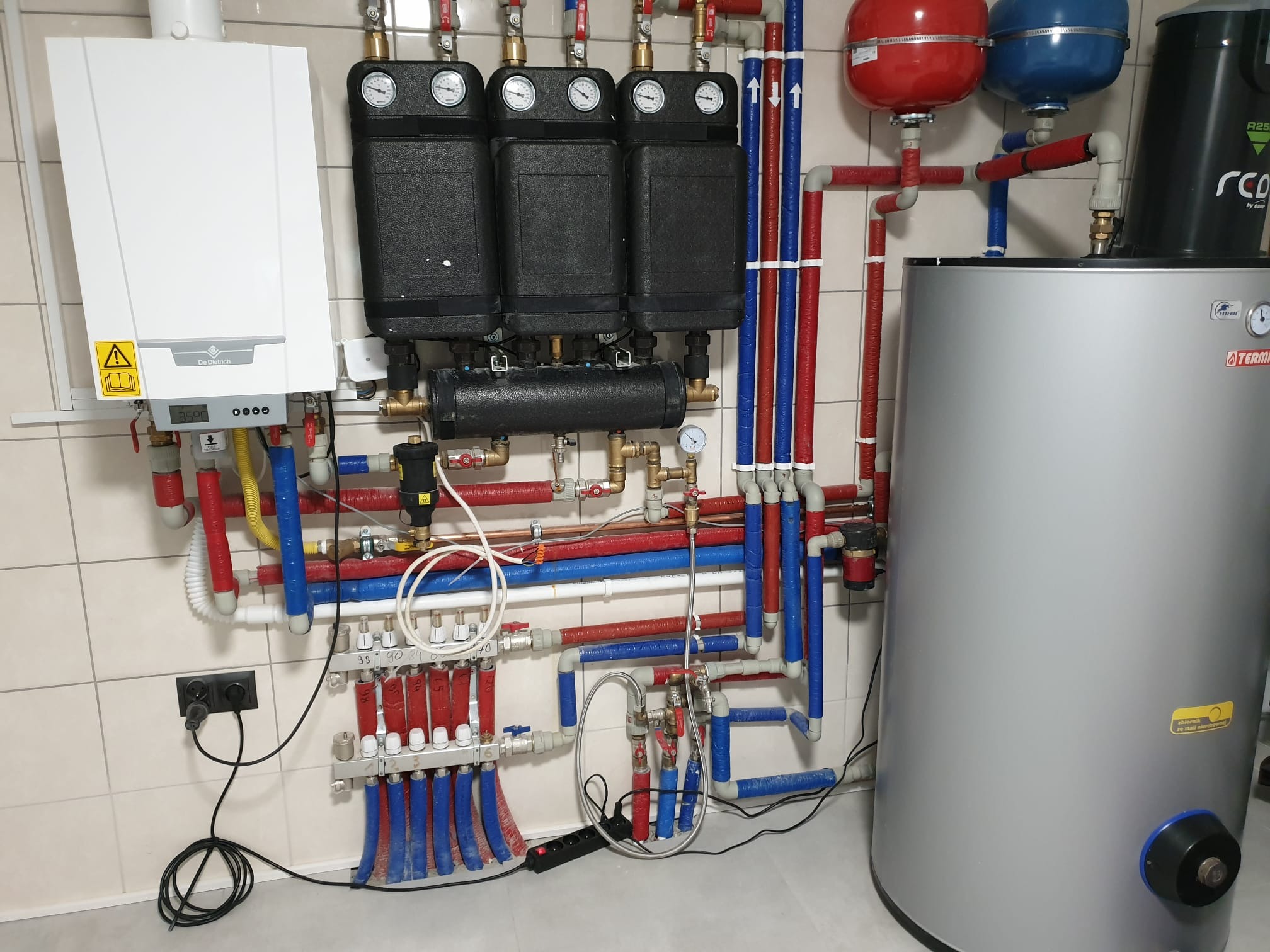 Kompleksowa instalacja grzewcza z kotłem gazowym, rozdzielaczami, zbiornikiem na wodę i licznymi rurami w kolorach czerwonym i niebieskim, zamontowana na kafelkowanej ścianie.