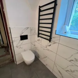 Nowoczesna łazienka po remoncie: białe płytki imitujące marmur, wisząca toaleta z czarnym przyciskiem spłukującym, czarny grzejnik drabinkowy, szara podłoga i okno z rolką papieru toaletowego...