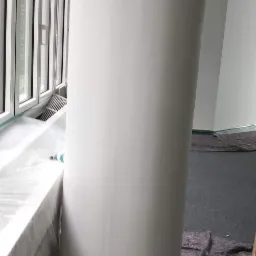 Pomalowany na biało okrągły filar w jasnym wnętrzu, zabezpieczone okno i podłoga folią malarską.