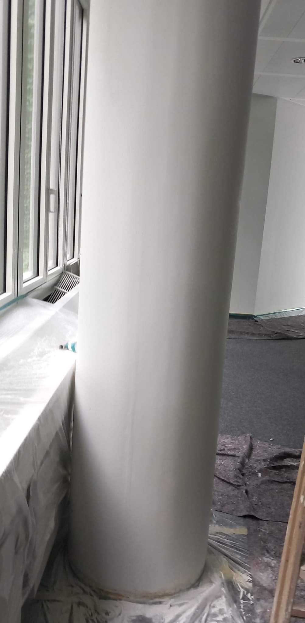 Pomalowany na biało okrągły filar w jasnym wnętrzu, zabezpieczone okno i podłoga folią malarską.