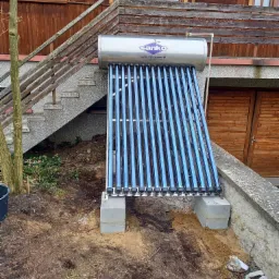 Kolektor słoneczny rurowy marki Sanko zamontowany na betonowych bloczkach przy drewnianym budynku z widocznymi schodami zewnętrznymi i fragmentami gruzu na ziemi.