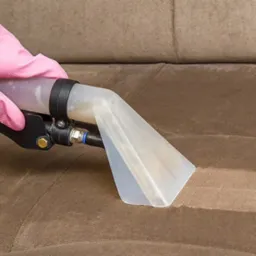 Beżowa kanapa czyszczona parowo-wodnym odkurzaczem, widoczna różnica w kolorze tapicerki po zabiegu, ręka w różowej rękawicy trzyma dyszę ssącą.