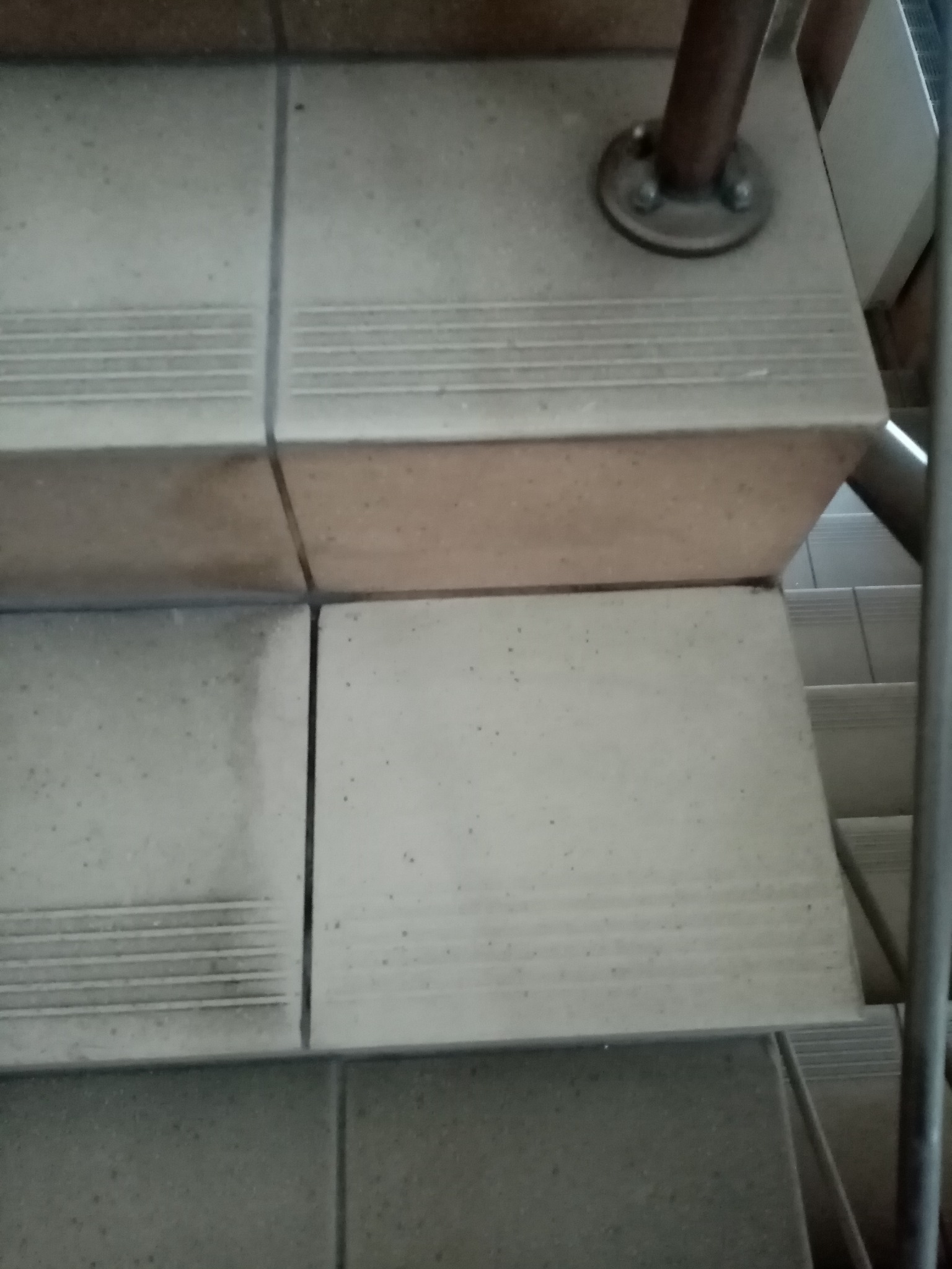 Szczegółowe ujęcie schodów wyłożonych płytkami ceramicznymi z antypoślizgowymi rowkami, widoczna metalowa poręcz oraz brud na powierzchni płytek.