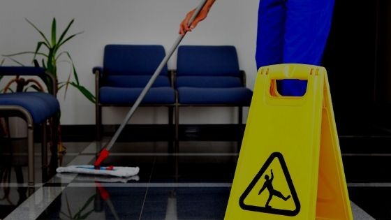 Osoba w niebieskim uniformie myje podłogę mopem w biurze, obok żółty znak ostrzegawczy, w tle ciemne krzesła i roślina doniczkowa.