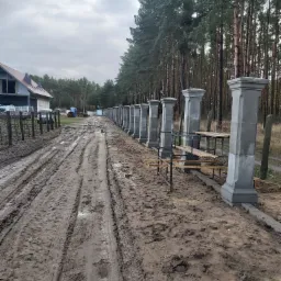 W trakcie budowy ogrodzenia z betonowych słupków, widoczne podpory i błotnista droga dojazdowa, w tle las i dom w stanie surowym.