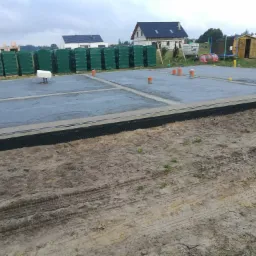 Przygotowany, zbrojony fundament pod budowę domu jednorodzinnego, zabezpieczony izolacją pionową, z widocznymi pionami kanalizacyjnymi i stosem materiałów budowlanych w tle.