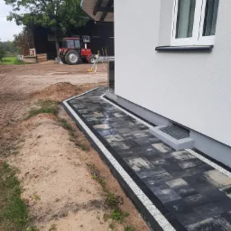 Ścieżka z ciemnej kostki brukowej z białym obramowaniem prowadząca wzdłuż ściany domu, w tle czerwony traktor i budynek gospodarczy.
