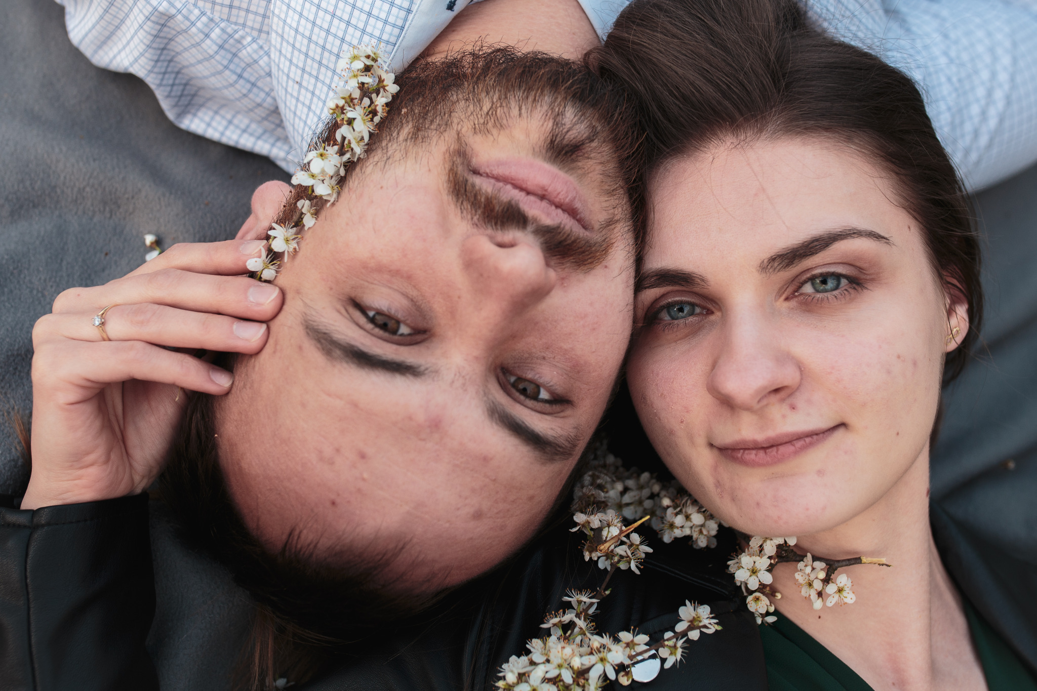 Zbliżenie na twarze młodej pary leżącej obok siebie, udekorowane gałązkami kwitnącej wiśni, z pierścionkiem zaręczynowym na palcu dłoni mężczyzny.