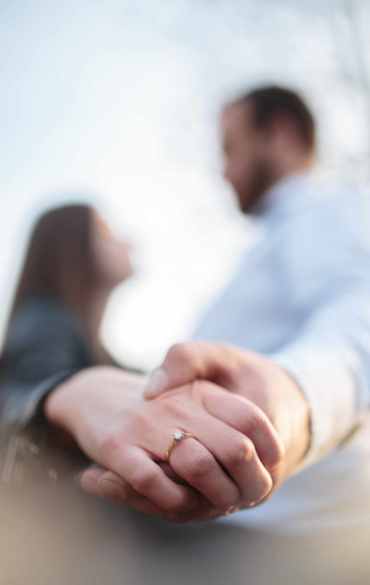 Złoty pierścionek zaręczynowy z pojedynczym brylantem na dłoni kobiety, delikatne rozmycie sylwetek pary w tle.