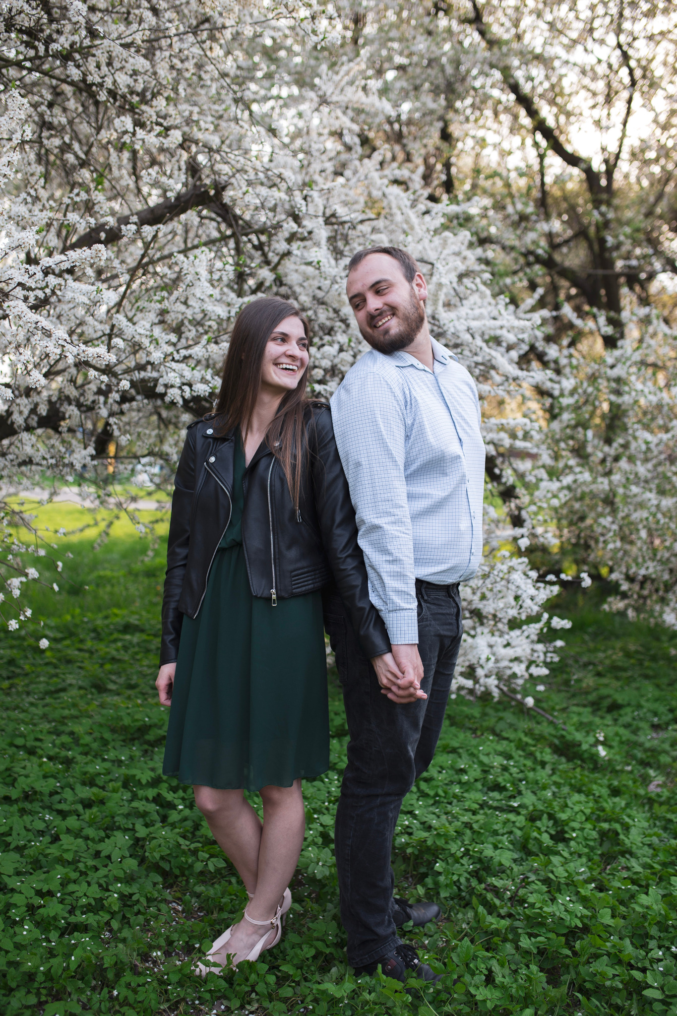 Uśmiechnięta para trzymająca się za ręce, oparta plecami o siebie na tle kwitnącego drzewa z białymi kwiatami i zielonej trawy.