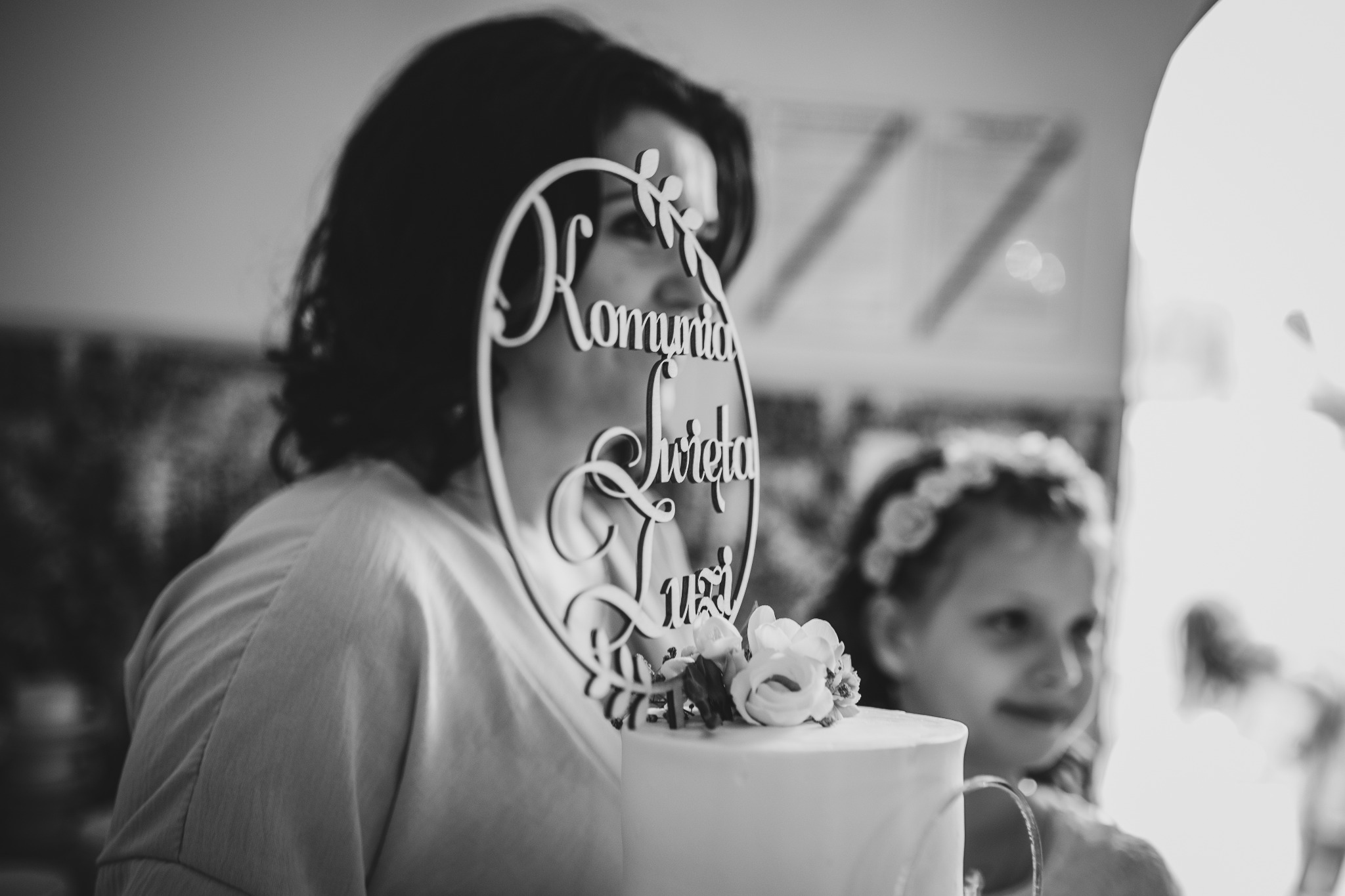 Czarno-białe zdjęcie tortu komunijnego z dekoracją 'Komunia Święta' i kwiatami, na tle uśmiechniętej mamy i dziewczynki w wianku.