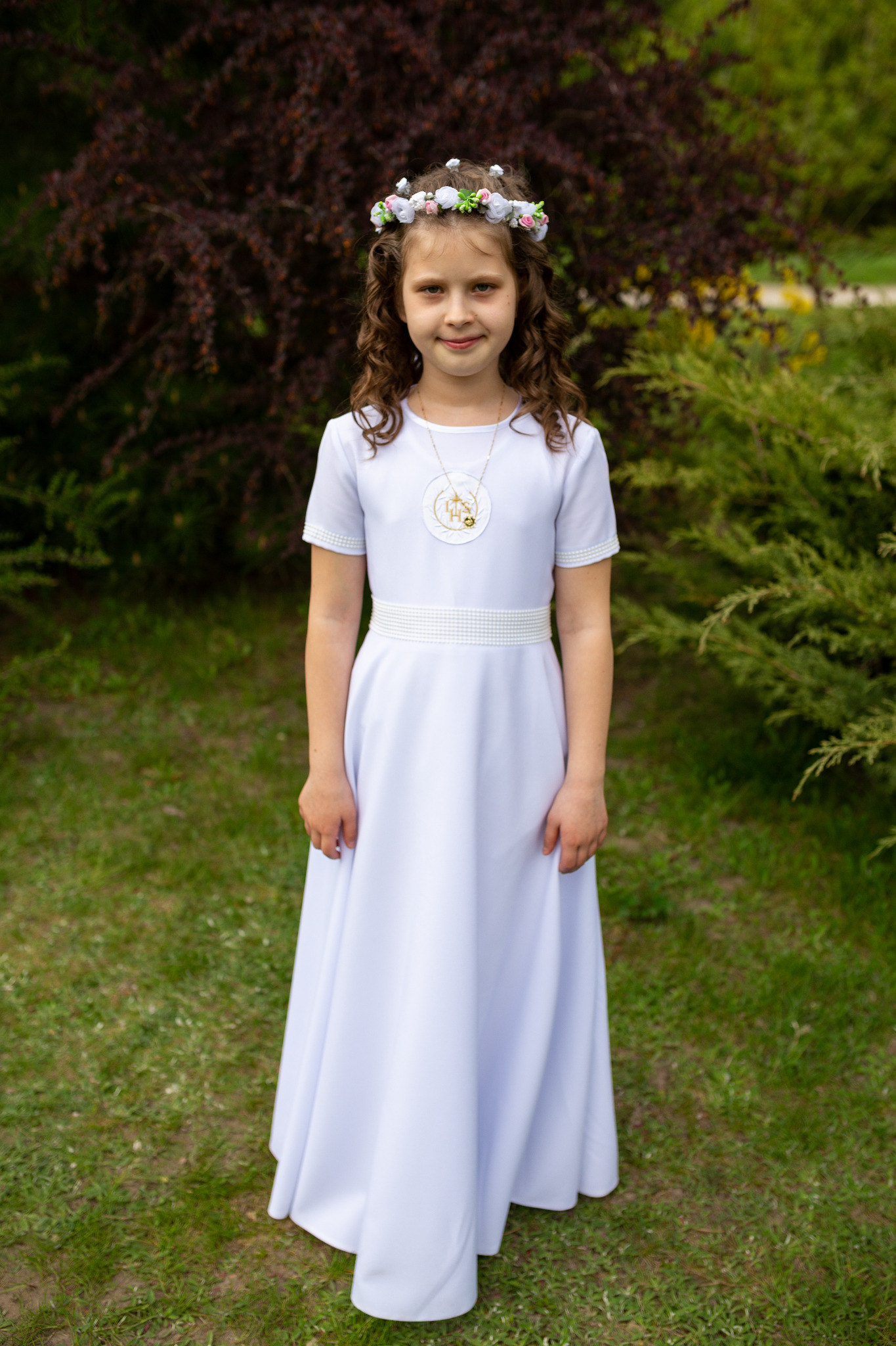 Dziewczynka w białej sukience komunijnej z wiankiem na głowie i medalikiem, stojąca na tle zieleni.