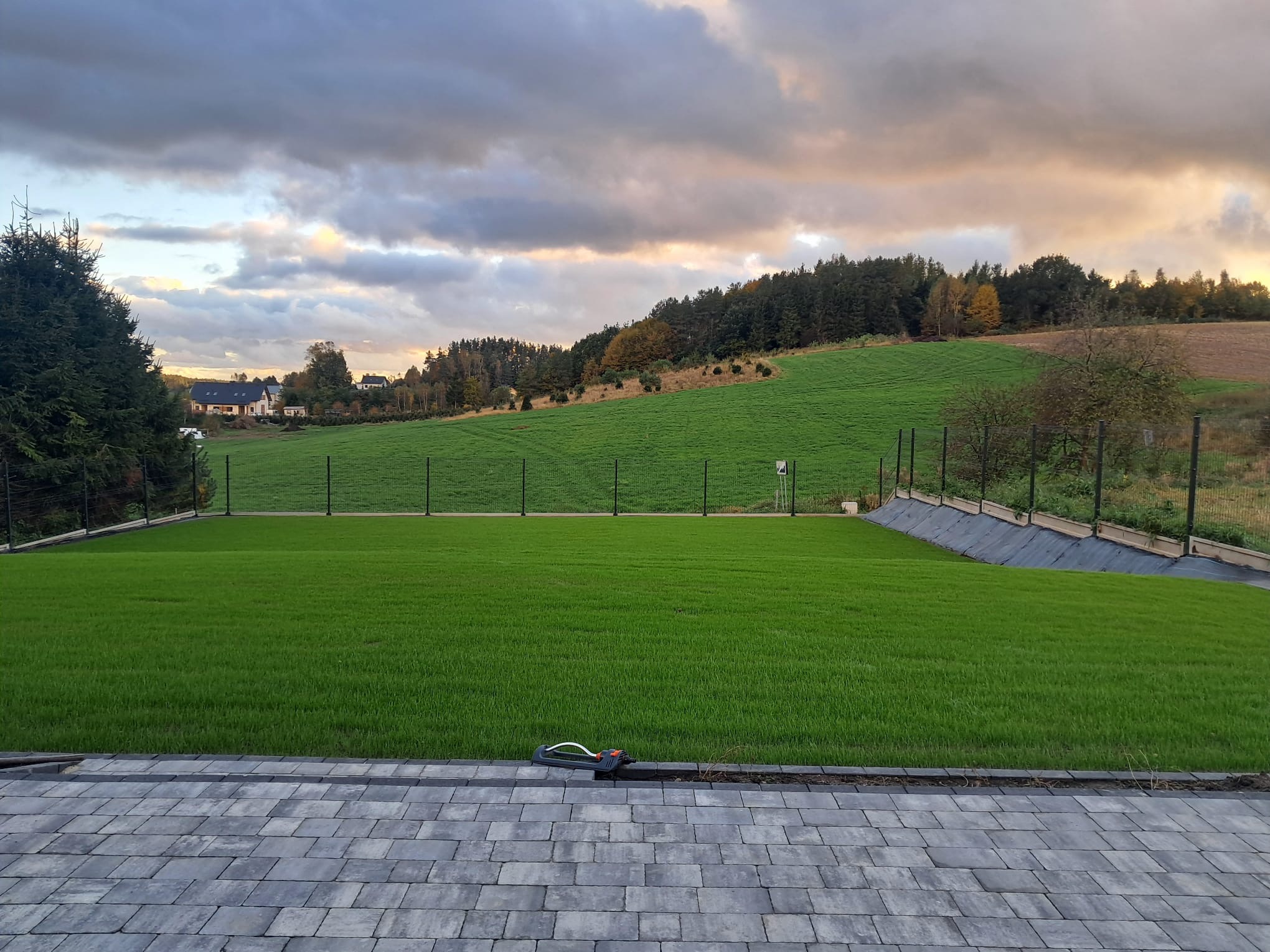 Widok na świeżo założony trawnik z systemem nawadniania, otoczony kostką brukową i siatką. W tle zielone wzgórza i las pod pochmurnym niebem o zachodzie słońca.