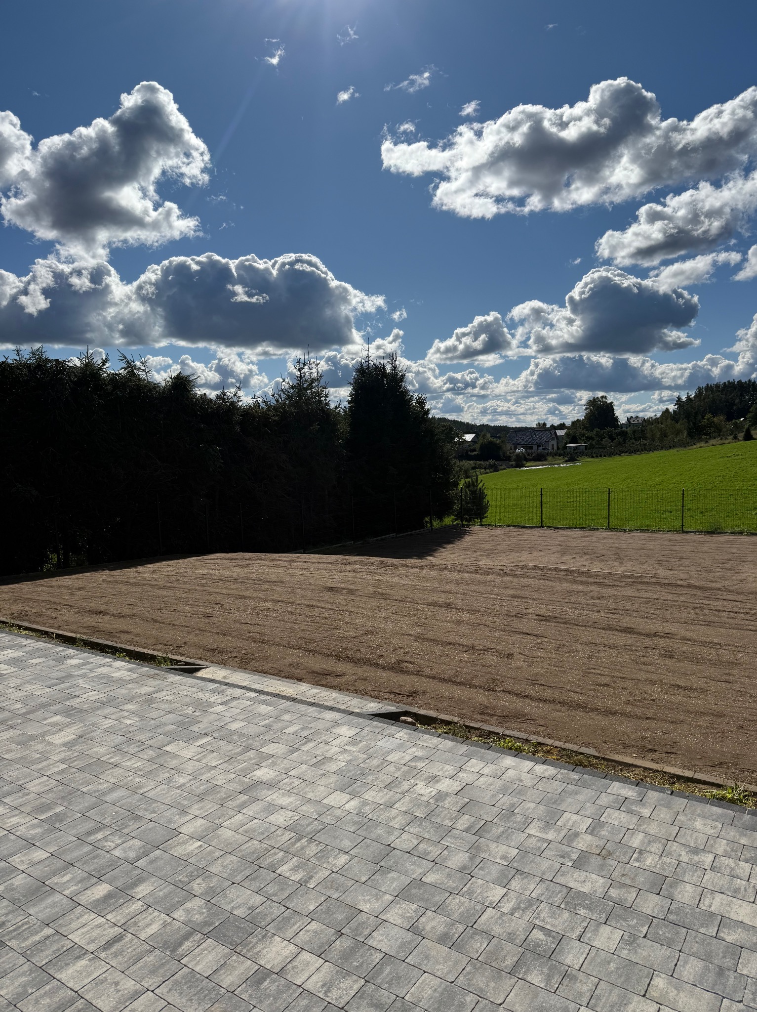 Wyrównany teren pod trawnik, widok z poziomu kostki brukowej. W tle zielone pole i las iglasty pod błękitnym niebem z chmurami.