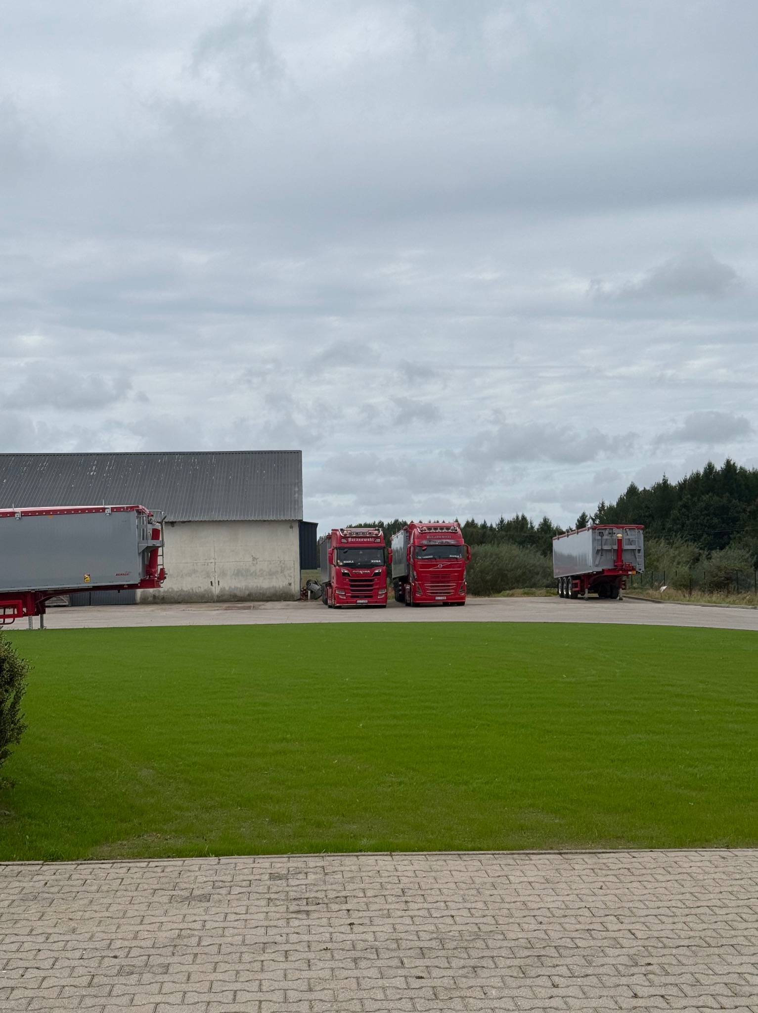 Dwa czerwone ciągniki siodłowe z naczepami na tle zielonego trawnika i szarego budynku. Pochmurne niebo. Widok z poziomu chodnika z kostki brukowej.
