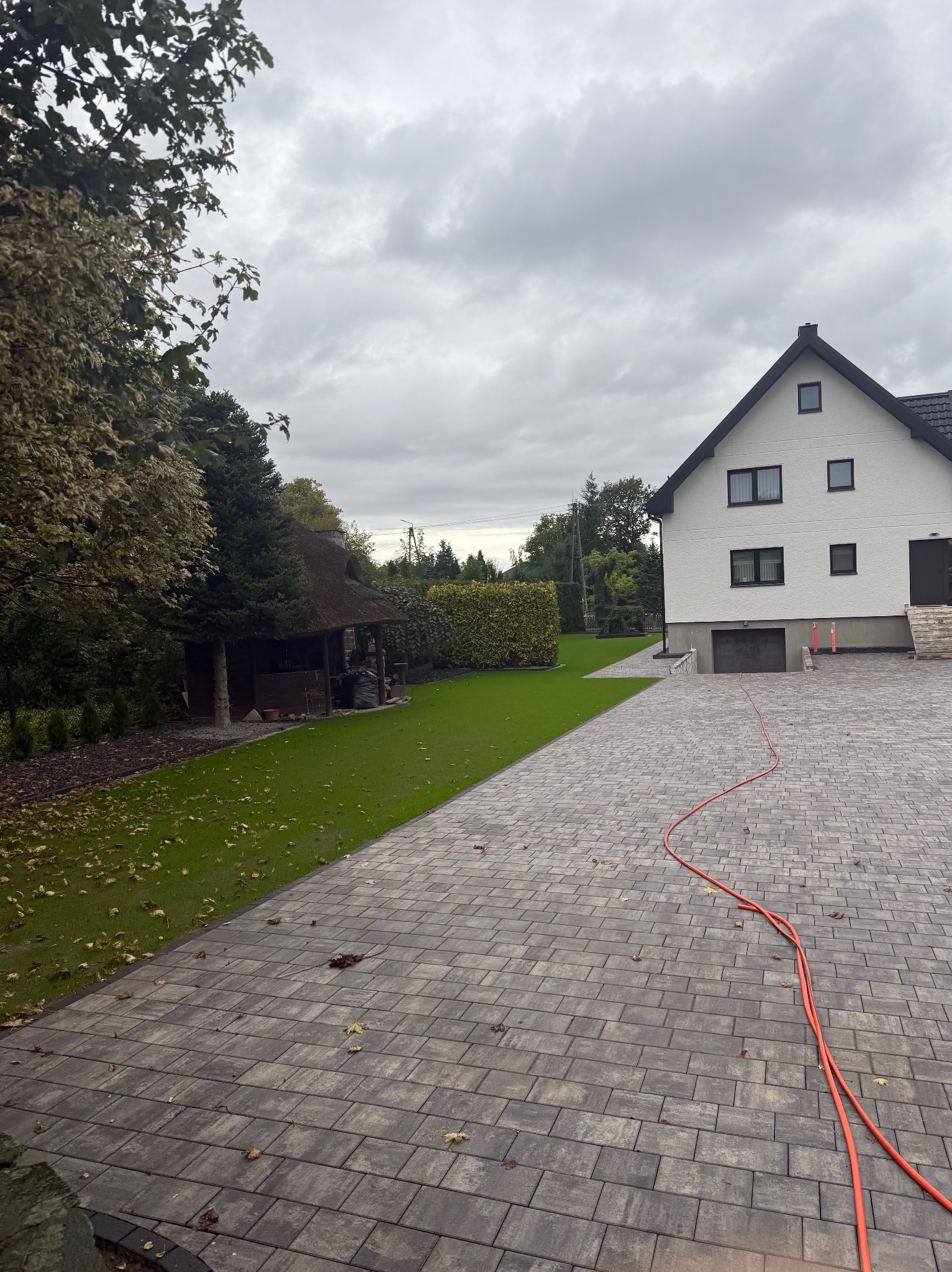 Nowoczesny dom z zadbanym trawnikiem i brukowanym podjazdem. Widoczna altana z trzcinowym dachem i pomarańczowy wąż na kostce brukowej. Pochmurne niebo.