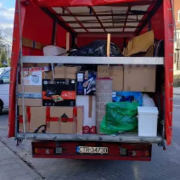 Otwarta przestrzeń ładunkowa czerwonego busa wypełniona kartonami, sprzętem AGD, dywanem i workami z dobytkiem, widok z tyłu na ulicę i fragment chodnika.