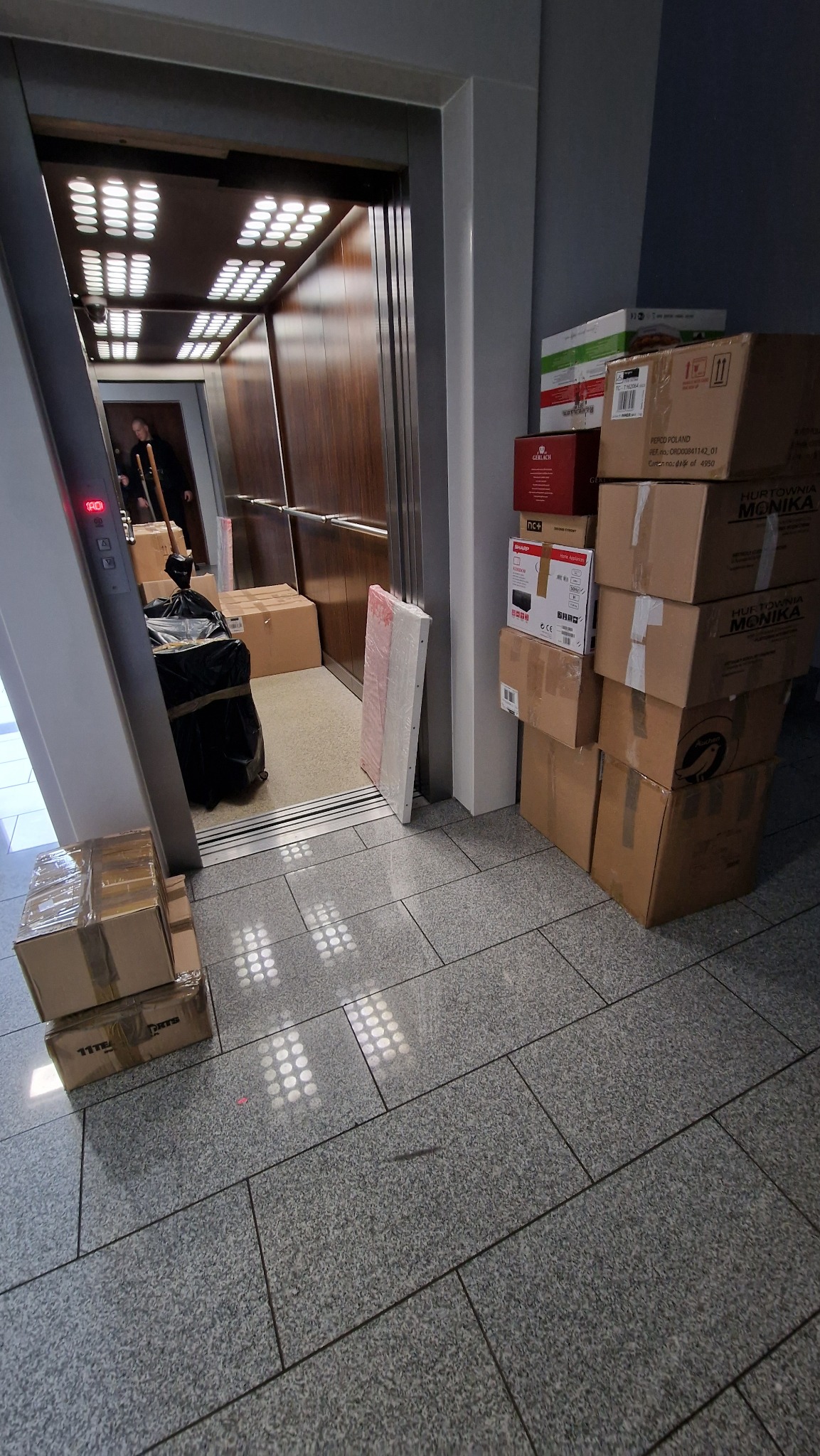 Kartonowe pudła i worki w windzie towarowej podczas przeprowadzki, widoczne oświetlenie sufitowe i fragment posadzki z płytek.