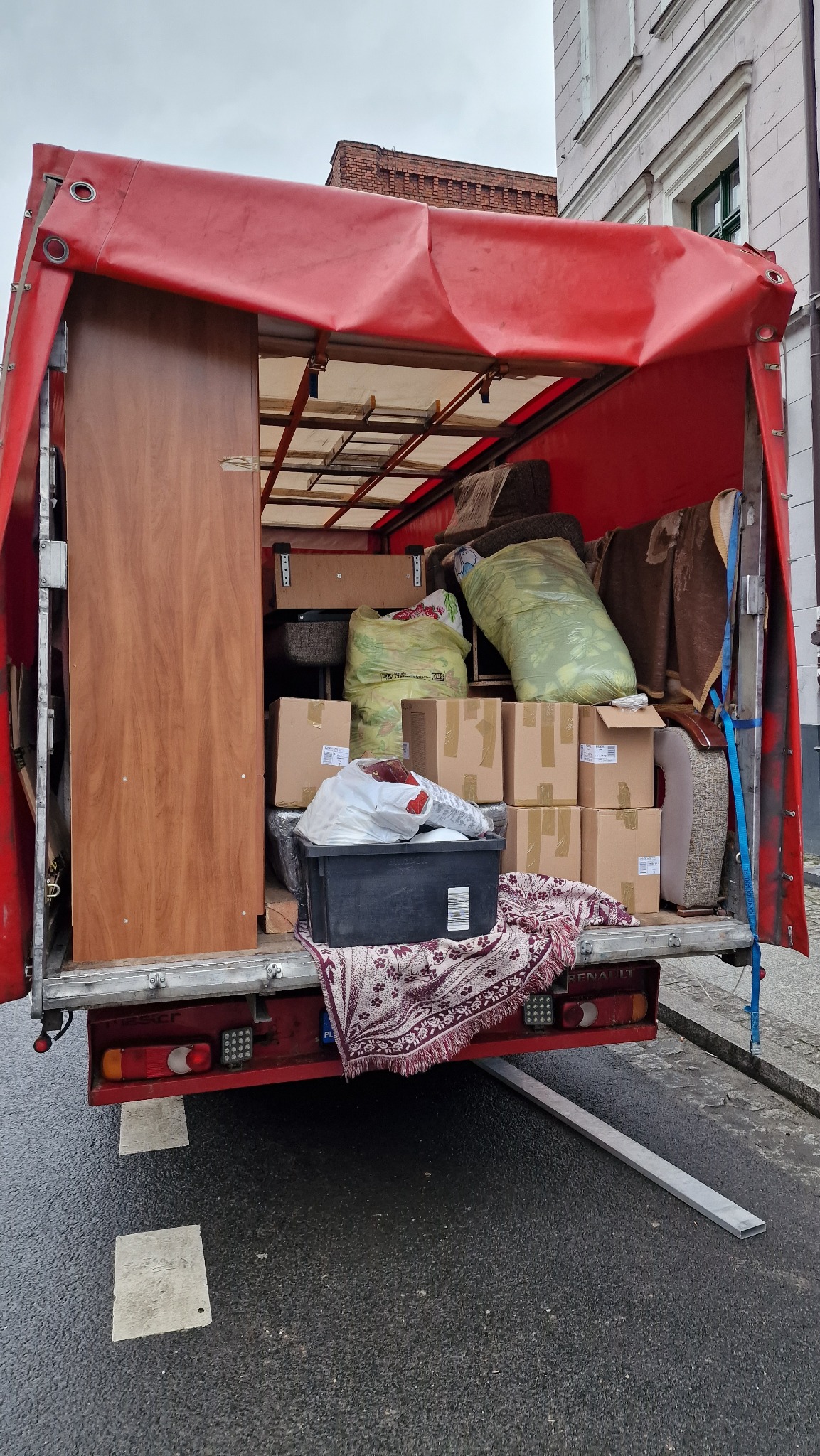 Wnętrze ciężarówki z czerwonym plandekowym dachem, załadowanej meblami, kartonami i workami podczas transportu, z rampą opuszczoną na mokrą nawierzchnię drogi.