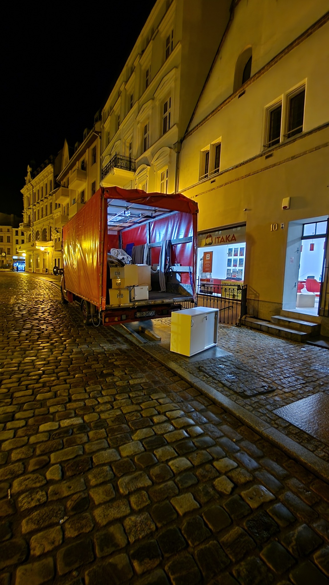Czerwony samochód dostawczy z otwartą przestrzenią ładunkową, wypełnioną kartonami i ubraniami, stoi na brukowanej ulicy w nocy, obok budynku z oświetlonym lokalem usługowym.