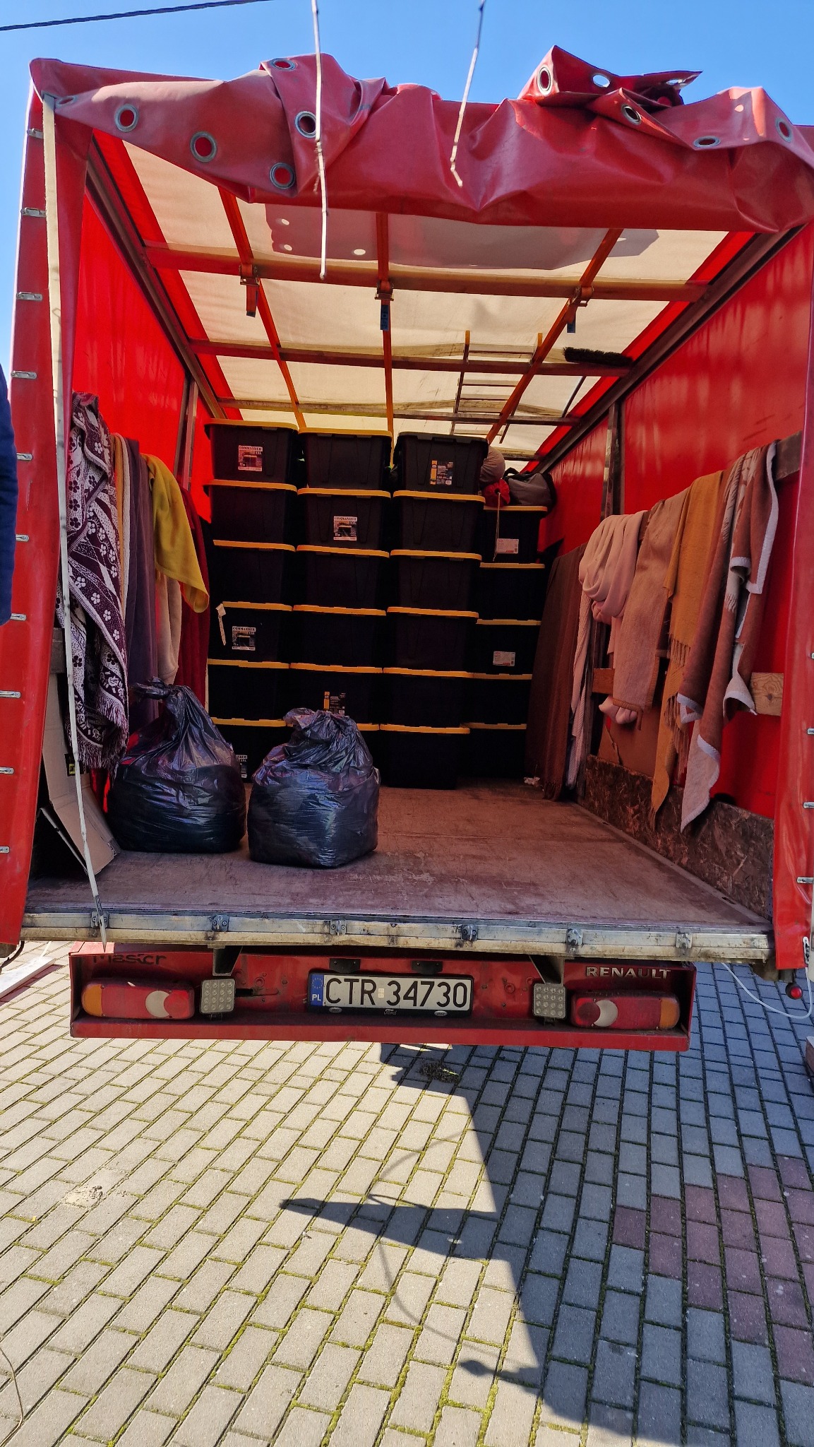 Wnętrze czerwonego busa z plandeką, wypełnione czarnymi pojemnikami z żółtymi elementami, workami na śmieci i ubraniami podczas przeprowadzki.