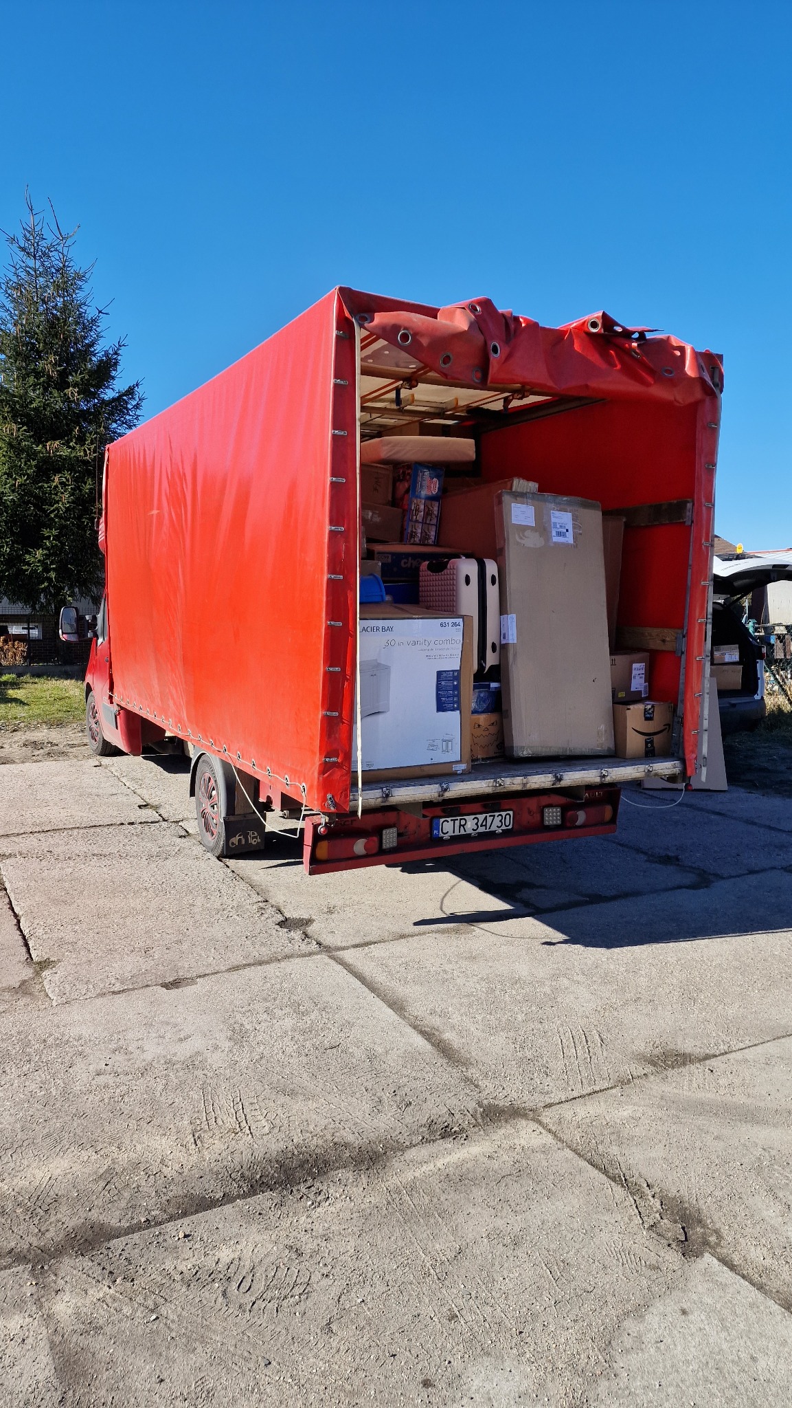 Czerwony samochód dostawczy z otwartą plandeką załadowany kartonami i meblami, widoczne tablice rejestracyjne CTR 34730.