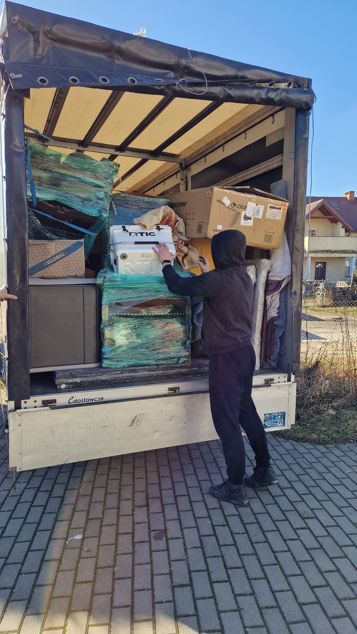 Osoba w czarnym stroju umieszcza białą lodówkę turystyczną na stercie zapakowanych przedmiotów wewnątrz otwartej przestrzeni ładunkowej ciężarówki z plandeką, widoczne kartony i meble zabezpieczone...