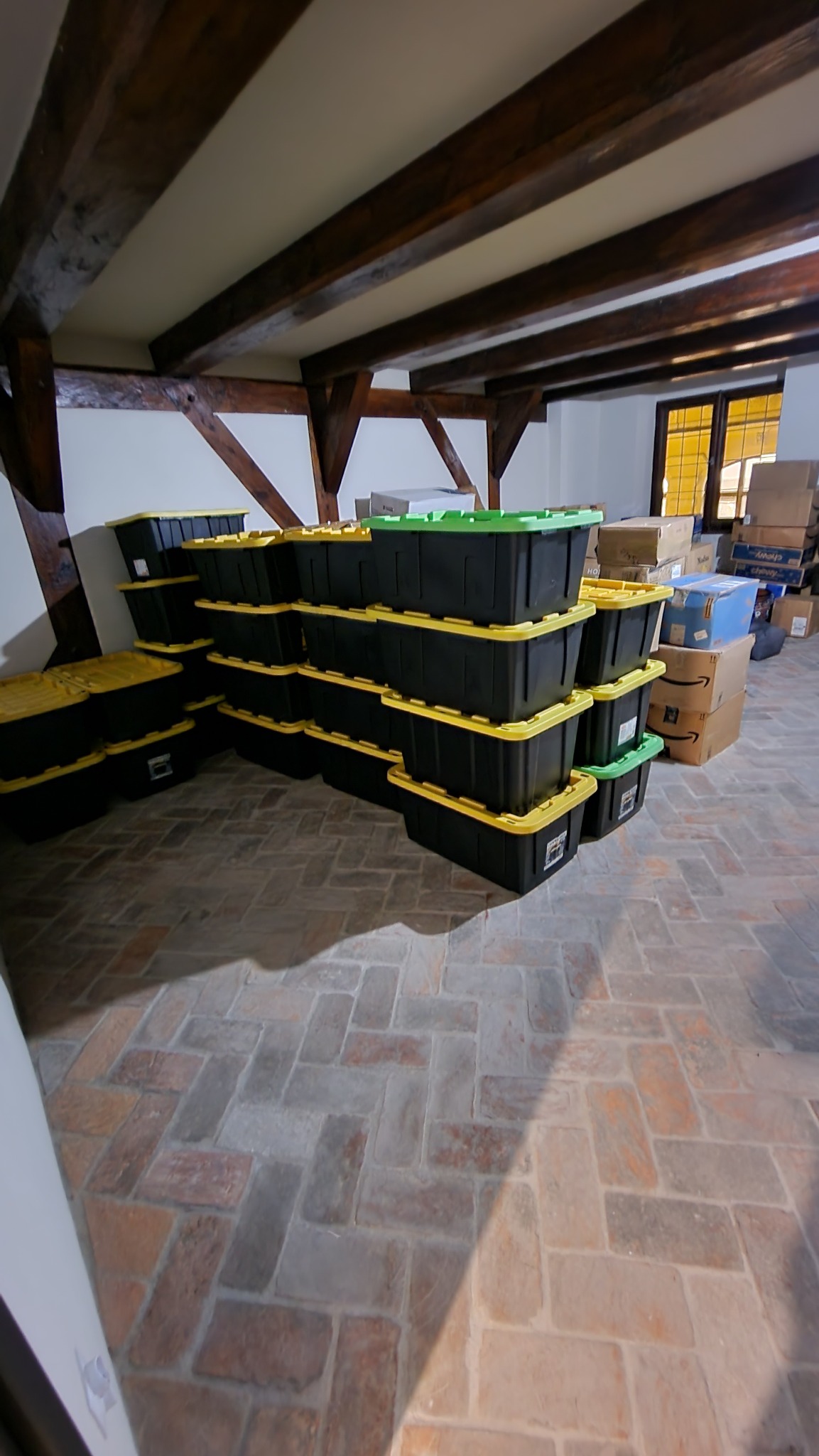Stos czarnych plastikowych pojemników z żółtymi i zielonymi pokrywami, ułożonych w pomieszczeniu z ceglaną podłogą i drewnianymi belkami stropowymi, obok kartonowych pudeł.
