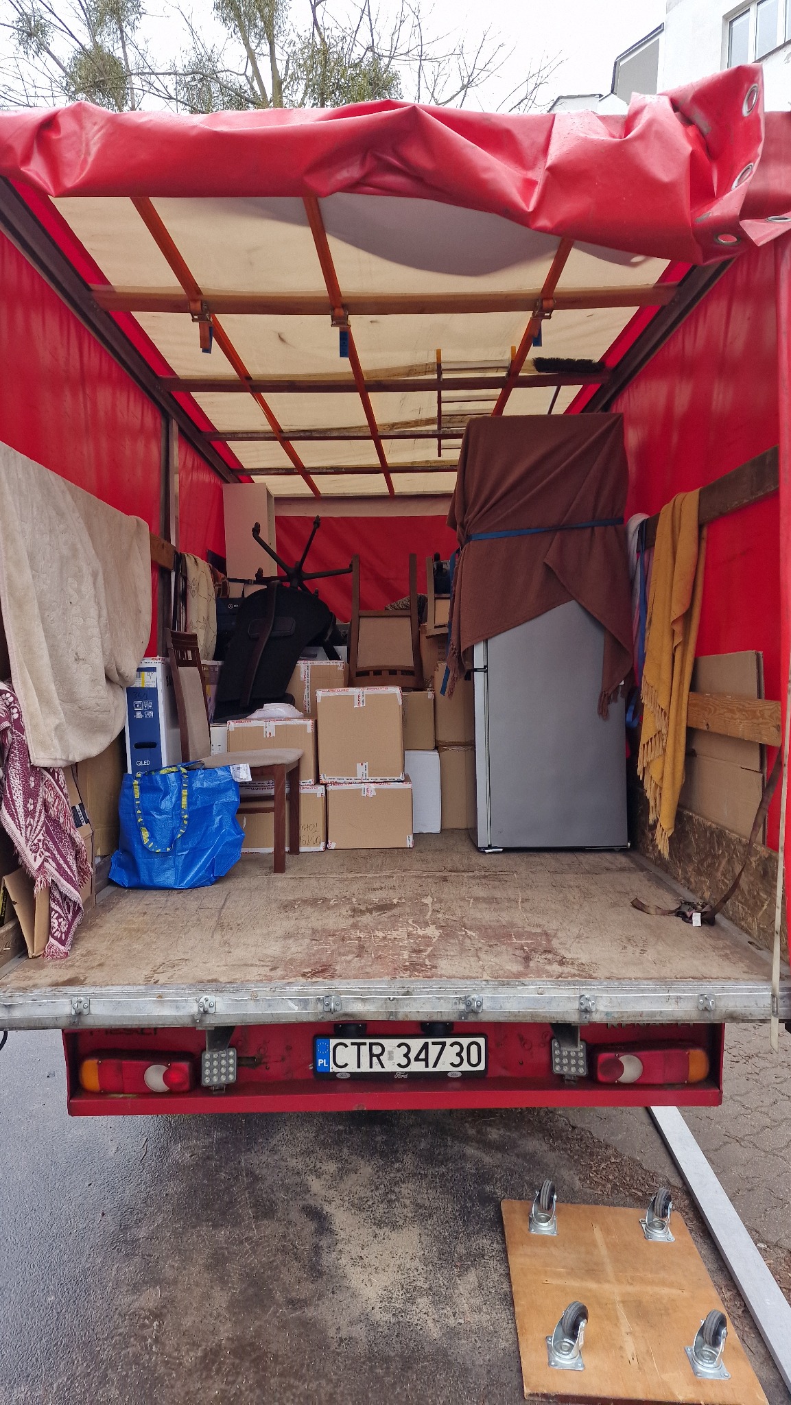 Wnętrze otwartej ciężarówki załadowanej kartonami, meblami i sprzętem AGD, zabezpieczonymi do transportu. Widoczna niebieska torba IKEA i tablica rejestracyjna pojazdu.
