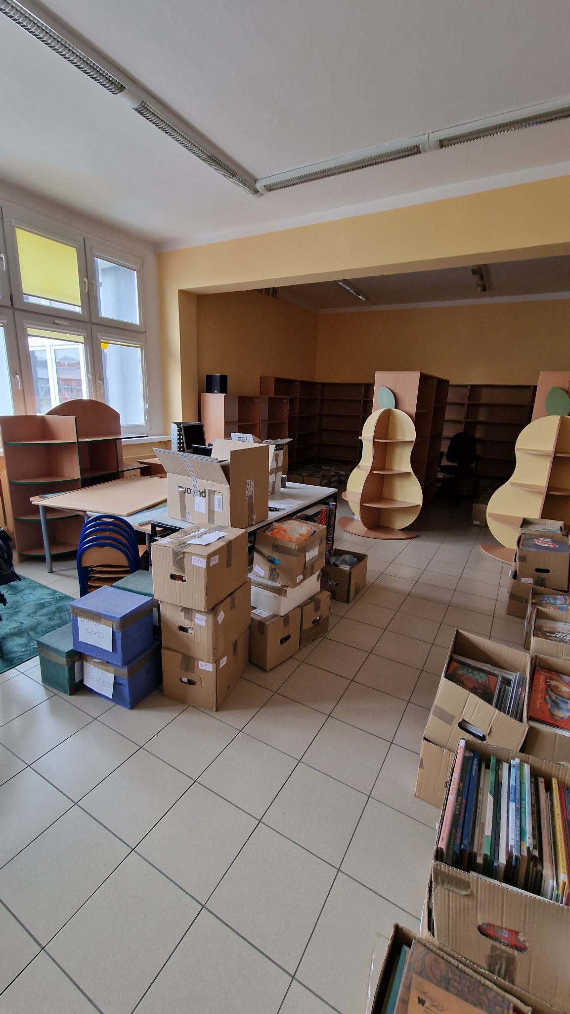 Kartonowe pudła i skrzynki z zawartością w opustoszałej sali lekcyjnej podczas przeprowadzki, widoczne regały w kształcie gruszek i biurka z krzesłami.