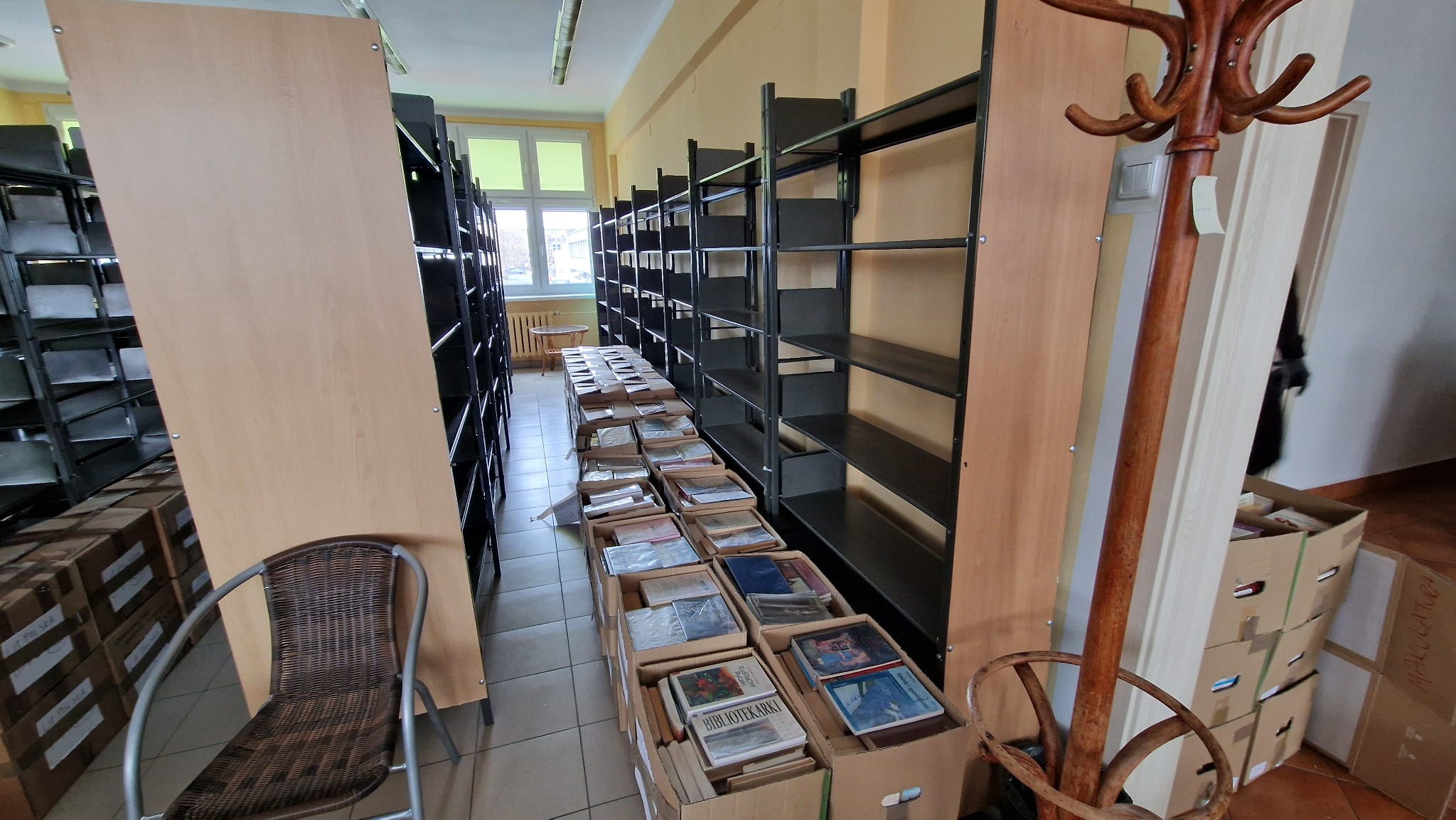 Korytarz biblioteki z metalowymi regałami, kartonami pełnymi książek, wiklinowym krzesłem i drewnianym wieszakiem na ubrania podczas pakowania przed przenosinami.