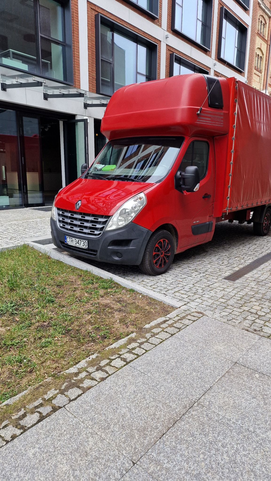 Czerwony samochód dostawczy Renault Master z plandeką, zaparkowany przed nowoczesnym budynkiem z dużymi oknami, widoczne felgi z czerwonym akcentem.