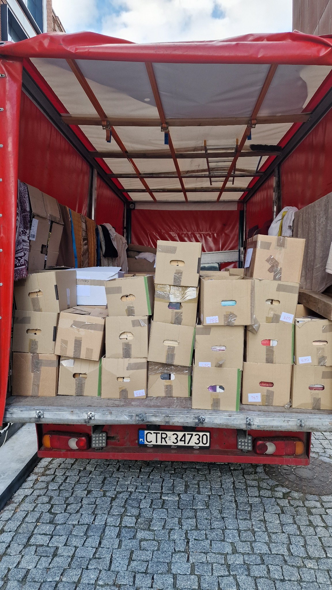 Wnętrze czerwonej ciężarówki wypełnionej kartonami z etykietami, widok z tyłu, tablica rejestracyjna CTR 34730 widoczna.