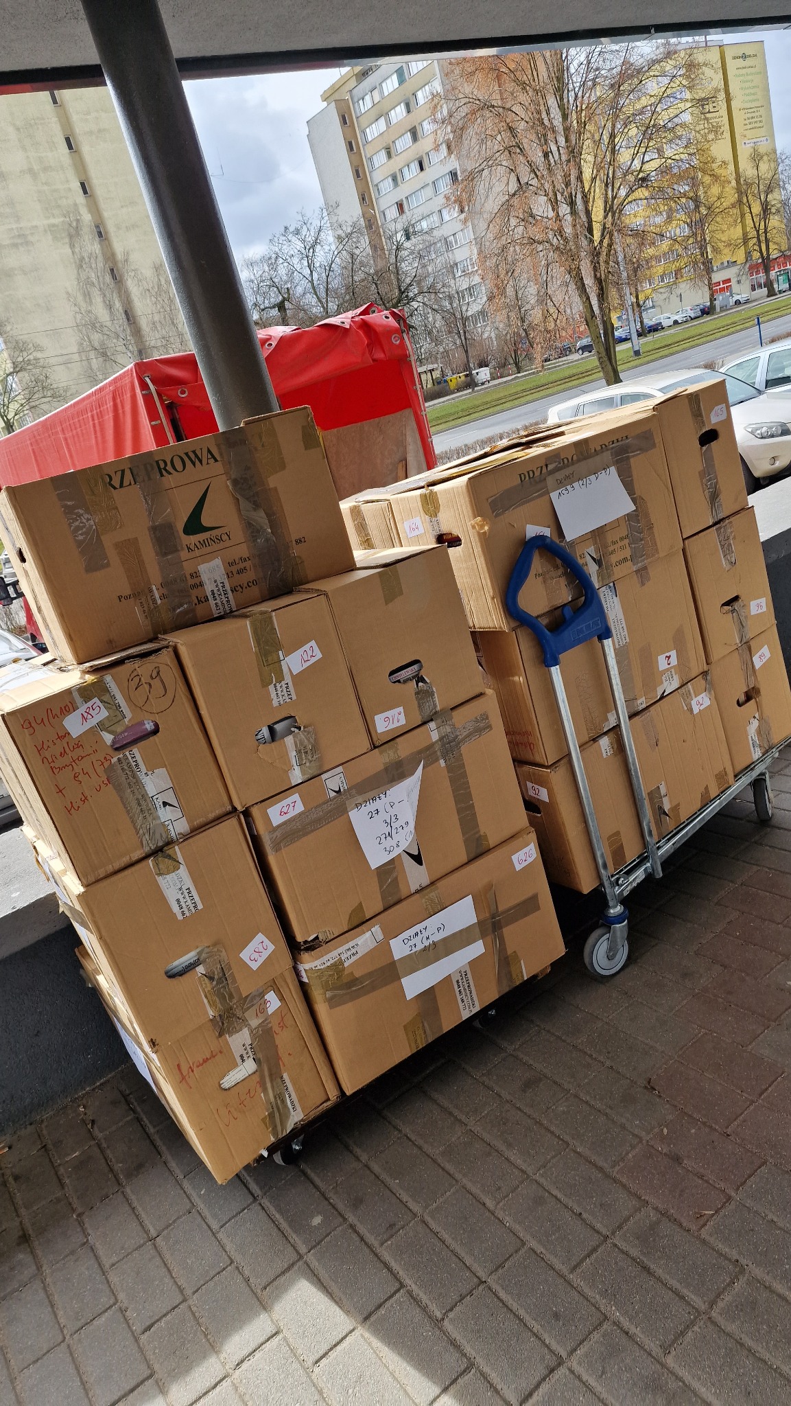 Kartonowe pudła z zawartością, zabezpieczone taśmą, ułożone na wózku transportowym z niebieską rączką, przed budynkiem mieszkalnym i zaparkowanymi samochodami.