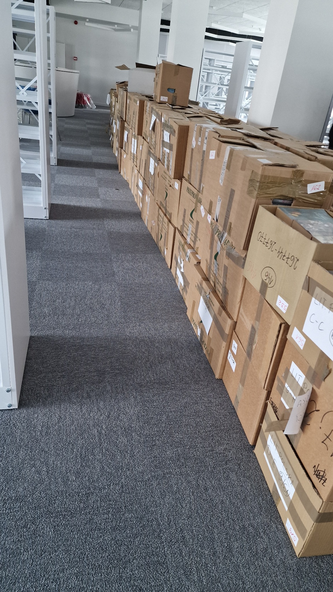 Rząd kartonowych pudeł zabezpieczonych taśmą, ustawionych wzdłuż ściany w jasnym pomieszczeniu biurowym z szarym dywanem, prawdopodobnie przygotowanych do transportu.