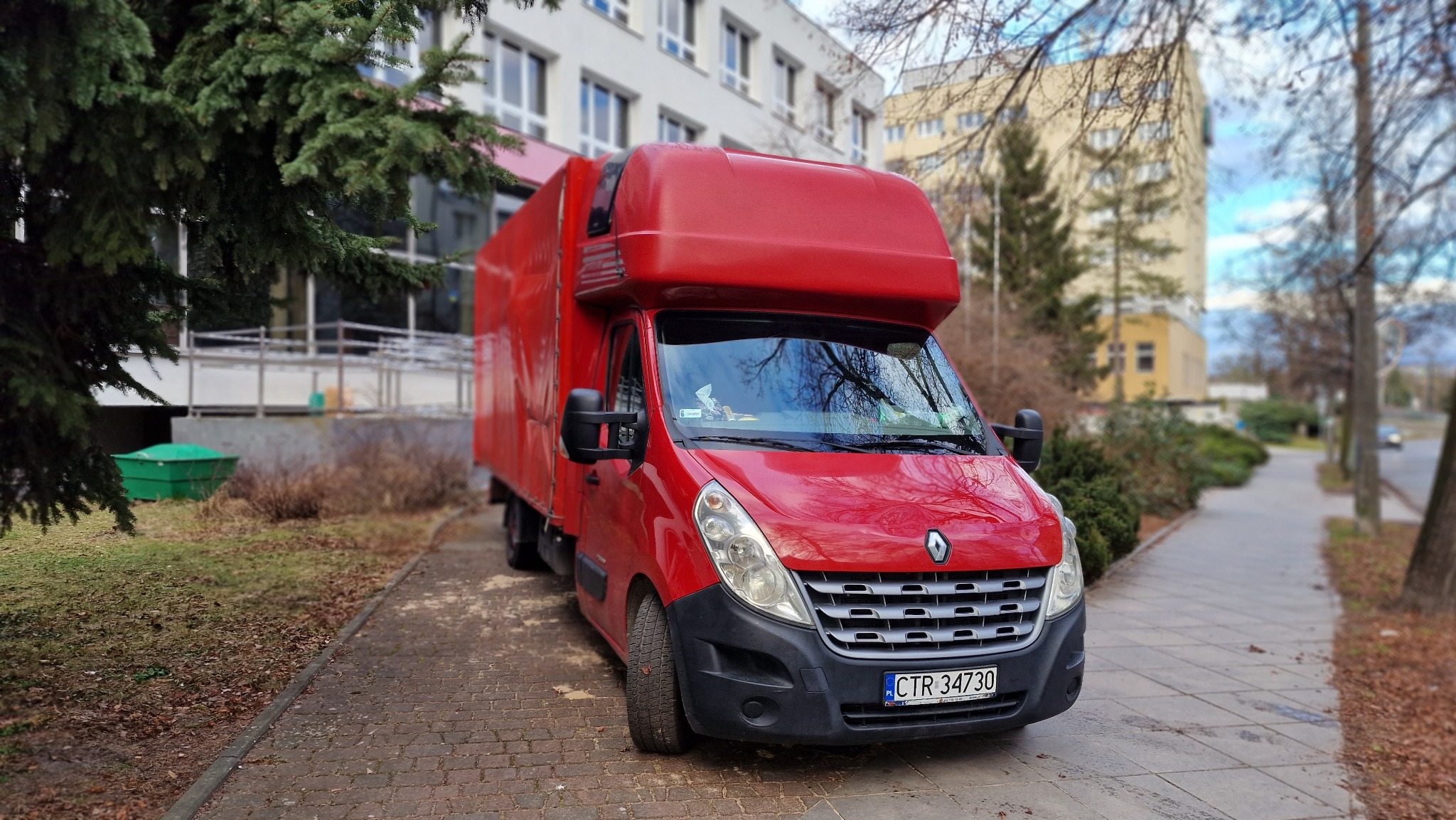 Czerwony samochód dostawczy Renault Master z zabudową kontenerową zaparkowany na chodniku w Toruniu, widoczne drzewa i budynki w tle.