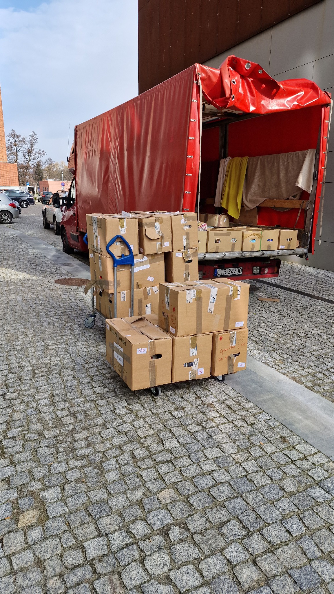 Stos kartonowych pudeł z niebieskim wózkiem transportowym przed otwartą przestrzenią ładunkową czerwonego samochodu dostawczego na brukowanym podłożu.