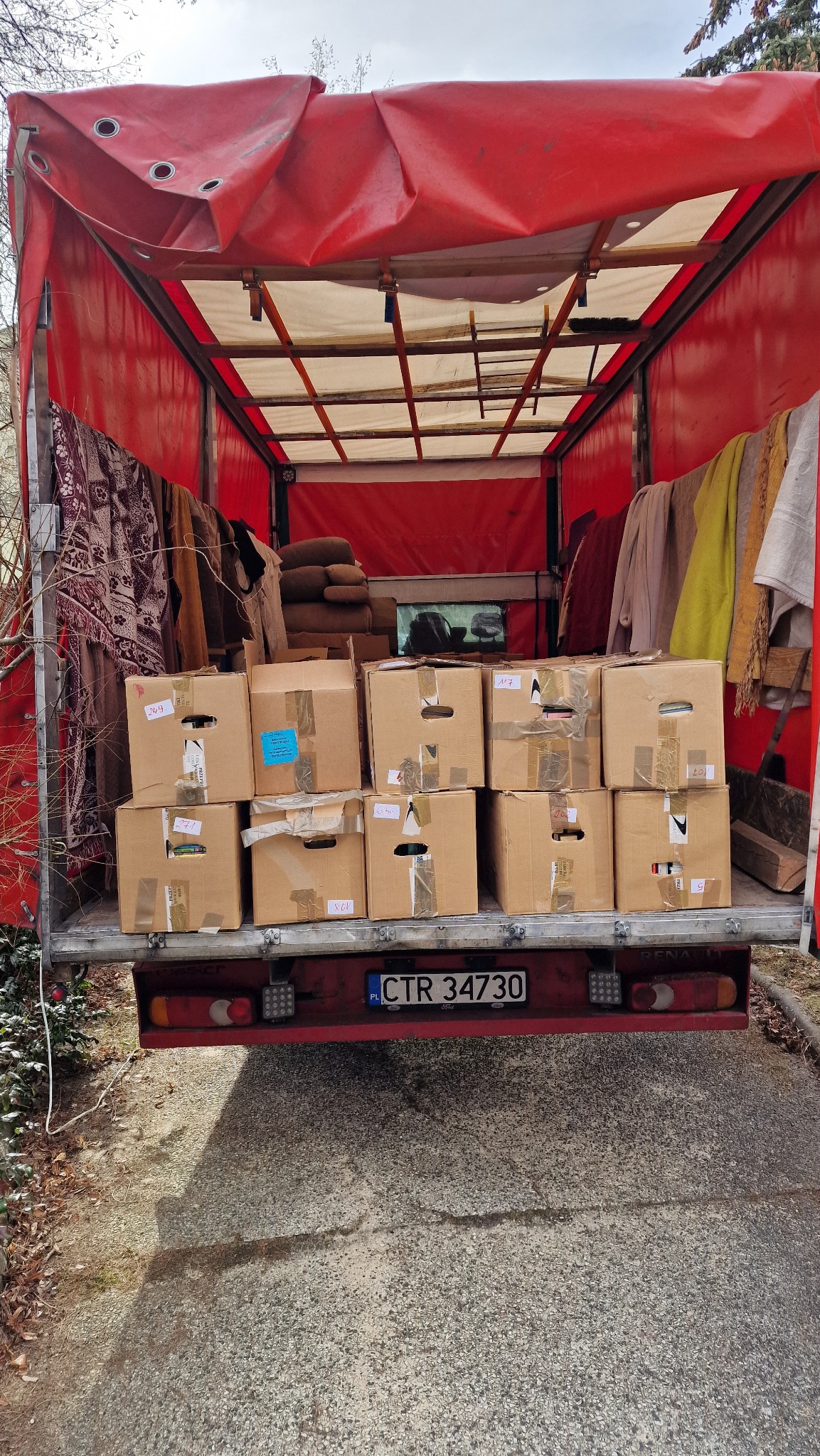 Kartonowe pudła z taśmą i numerami, załadowane do wnętrza czerwonej ciężarówki z plandeką, widok od tyłu z widoczną tablicą rejestracyjną.