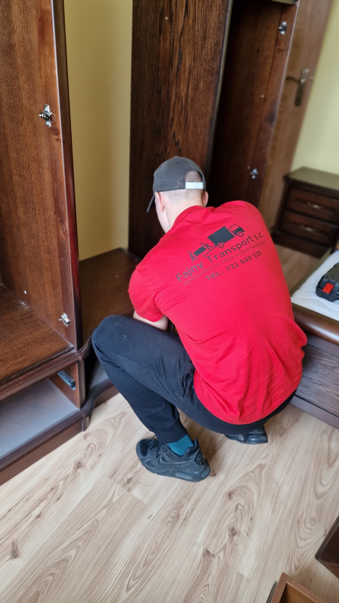 Mężczyzna w czerwonej koszulce z logo firmy transportowej demontuje drewnianą szafę podczas relokacji mebli.