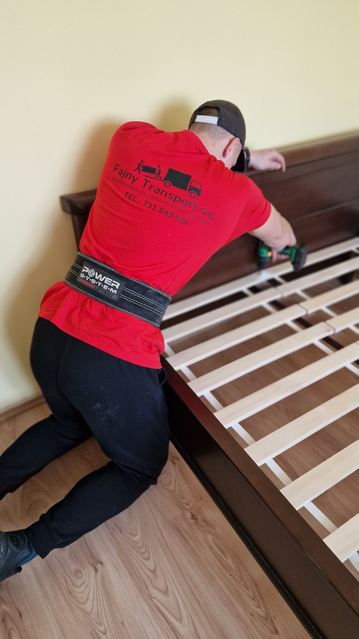 Pracownik w czerwonej koszulce firmy transportowej 'Fajny Transport' montuje drewniane łóżko za pomocą wkrętarki, zabezpieczony pasem Power System.
