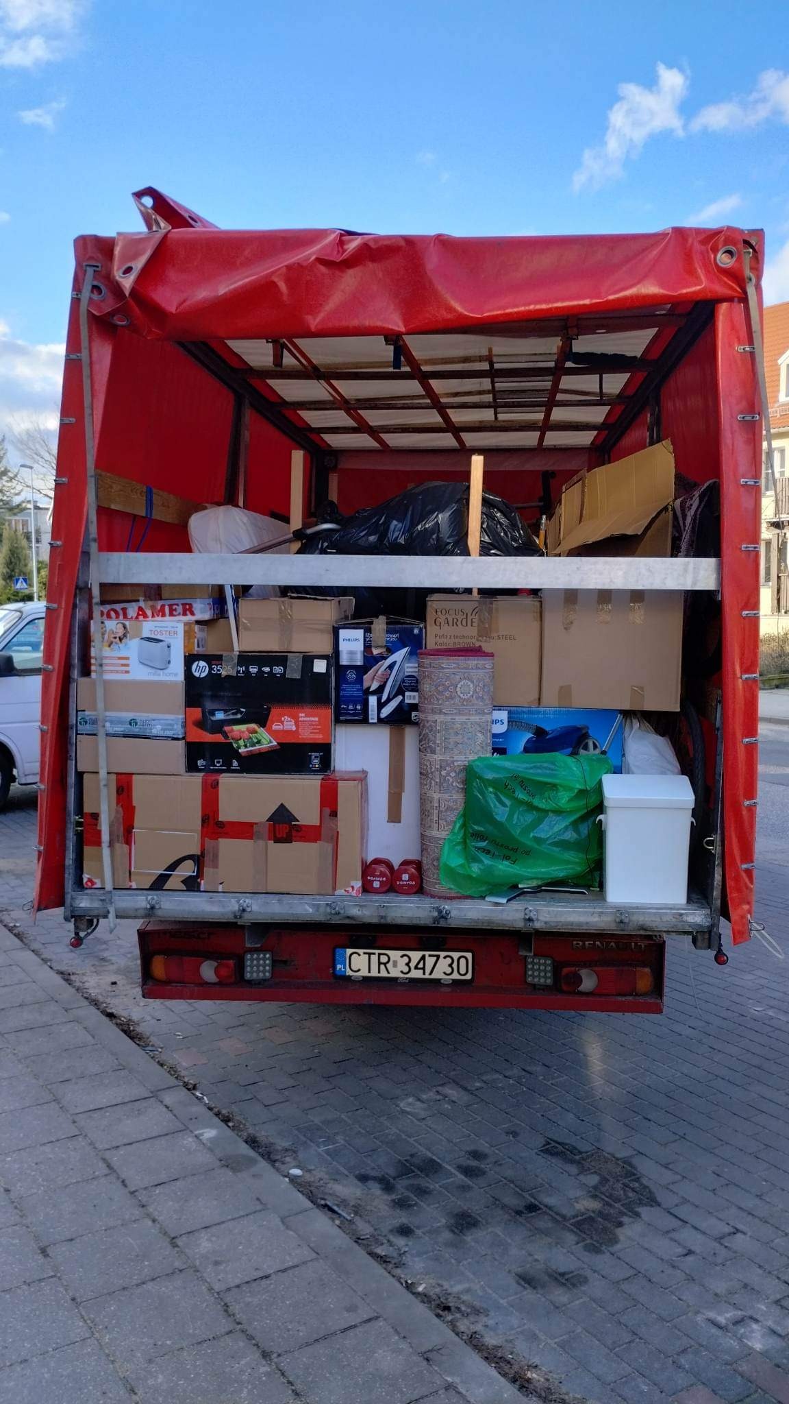 Otwarta przestrzeń ładunkowa czerwonego busa wypełniona kartonami, sprzętem AGD, dywanem i workami z dobytkiem, widok z tyłu na ulicę i fragment chodnika.
