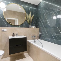 Pw Invest Home - Nowoczesna łazienka z czarnymi marmurowymi płytkami, okrągłym lustrem w złotej ramie, białą wanną i toaletą, oraz czarną szafką pod umywalką, oświetlona światłem LED.