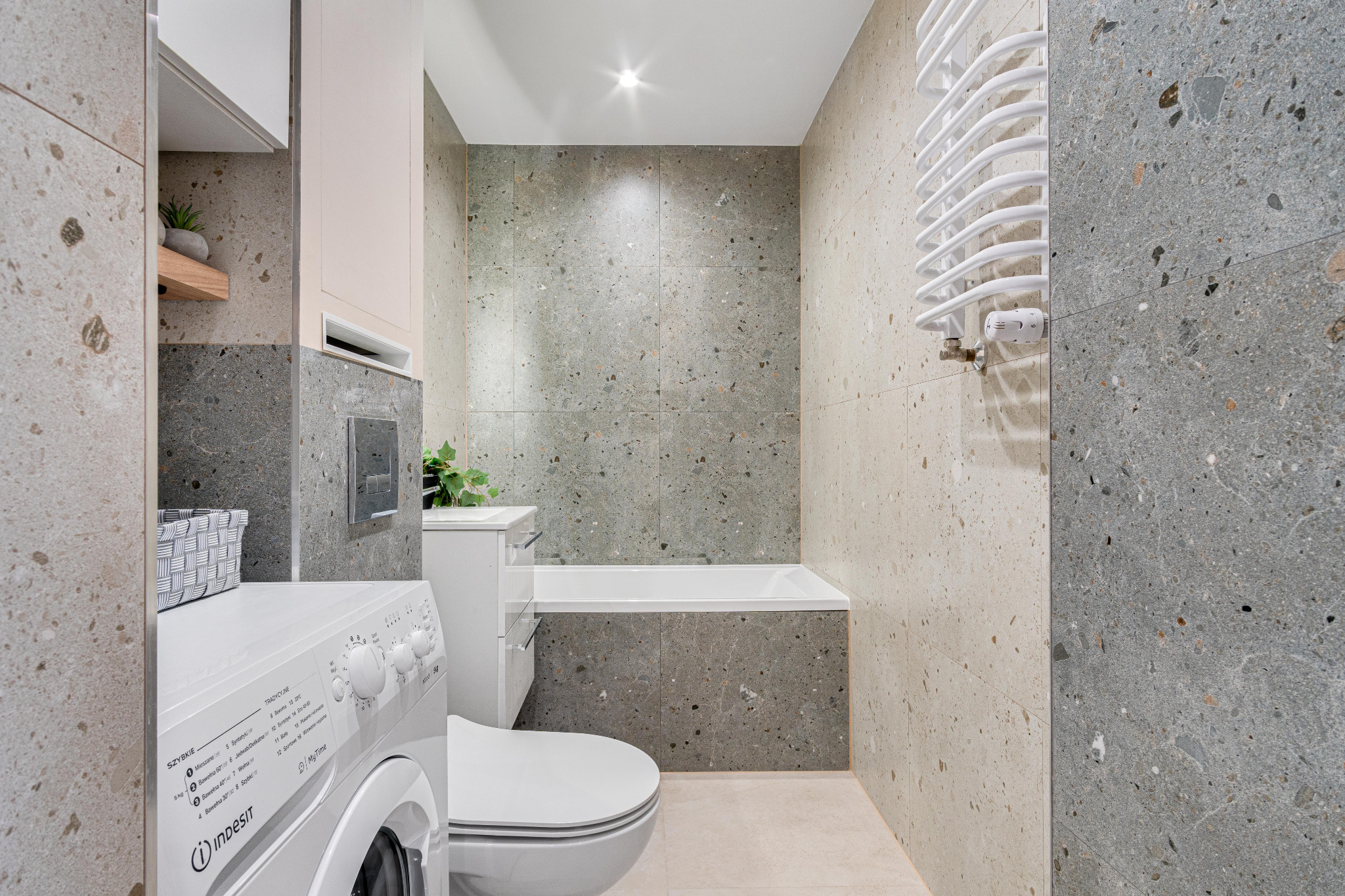 Nowoczesna łazienka z szarymi płytkami lastryko, białą wanną, toaletą i pralką Indesit, uzupełniona grzejnikiem drabinkowym i roślinami doniczkowymi.
