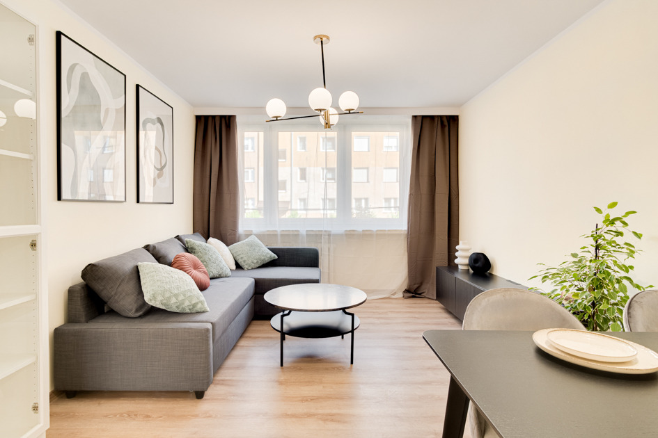 Jasne wnętrze mieszkania z szarą narożną sofą, okrągłym stolikiem kawowym, zasłonami w kolorze brązowym i nowoczesną lampą sufitową na tle okna z widokiem na budynek mieszkalny.