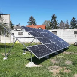 Panel słoneczny na metalowej konstrukcji, zamontowany na betonowych podstawach, na tle budynku z białej cegły i zielonego trawnika z żółtymi kwiatami, w słoneczny dzień.