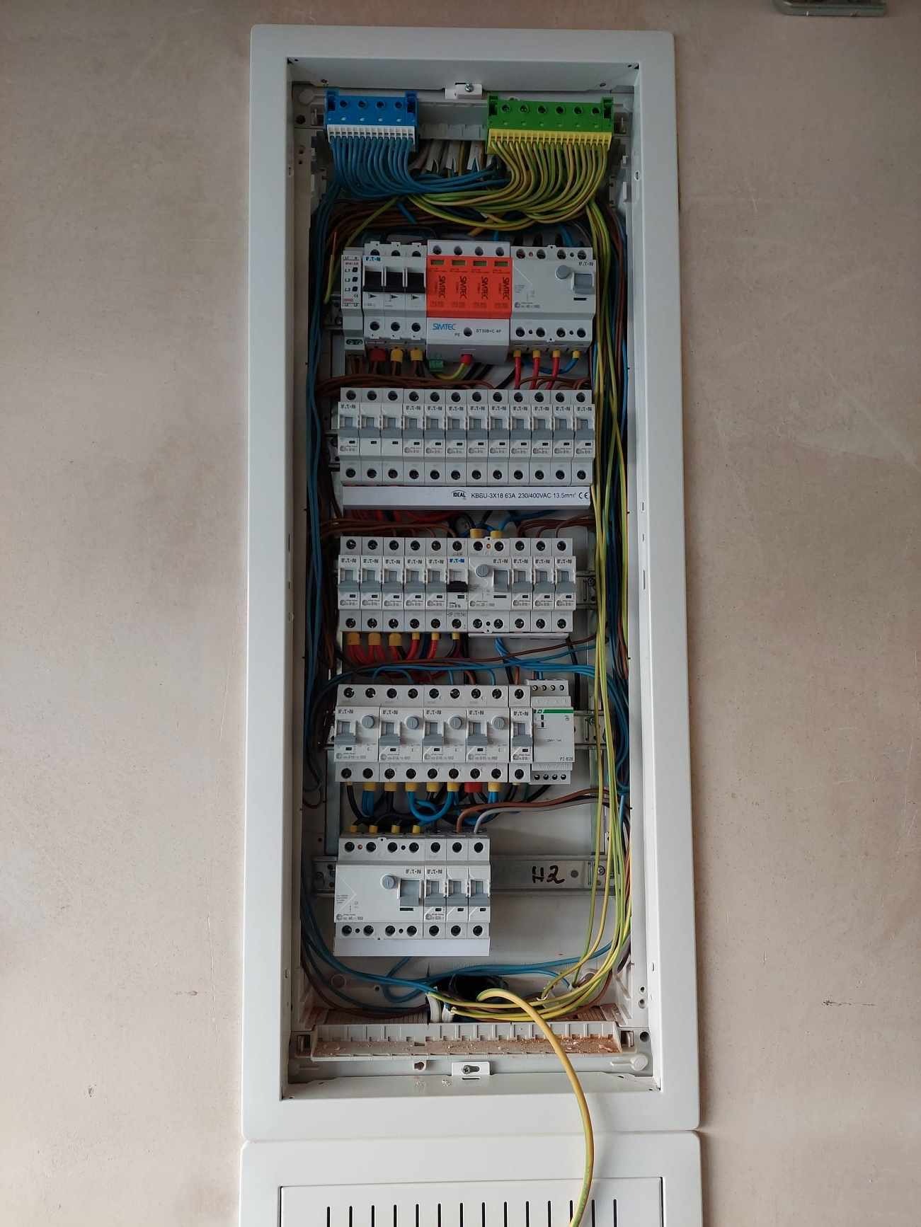 Otwarta skrzynka rozdzielcza z bezpiecznikami, widoczne okablowanie, listwy zaciskowe i wyłączniki różnicowoprądowe. Widoczne oznaczenia producentów: EATON, SIMTEC i IDEAL. Na ścianie widoczne...