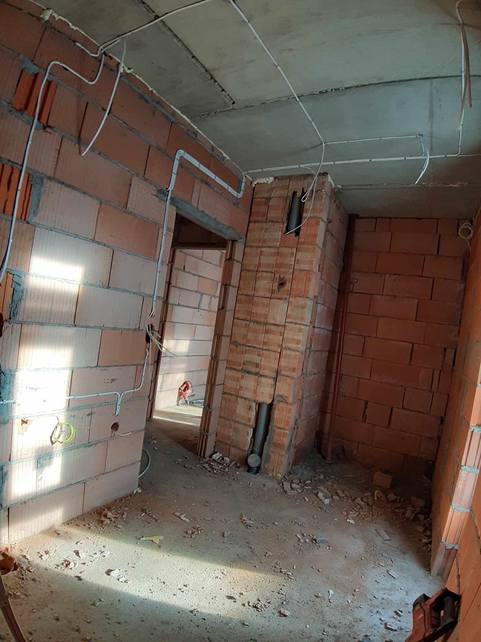 Surowy stan wnętrza z niedokończonymi ścianami z cegły, widoczne przewody elektryczne prowadzone po ścianach i suficie, oraz pion kanalizacyjny w narożniku.