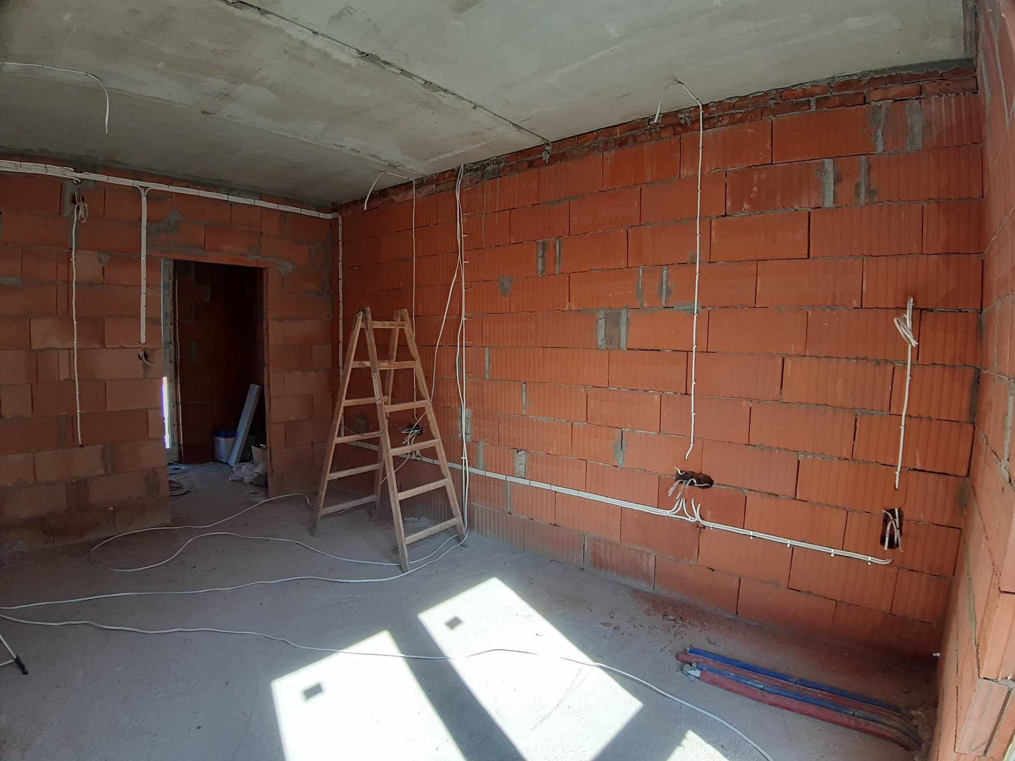 Wnętrze w trakcie instalacji elektrycznej: widoczne przewody w peszlach, puszki elektryczne w ścianach z czerwonej cegły, drewniana drabina i surowy betonowy strop.