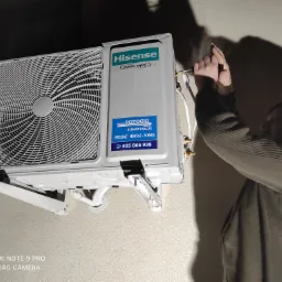 Instalacja klimatyzatora Hisense na ścianie. Widoczny monter w trakcie podłączania przewodów elektrycznych. Na urządzeniu naklejka z nazwą firmy Potocki Klimatyzacje i numerem telefonu.